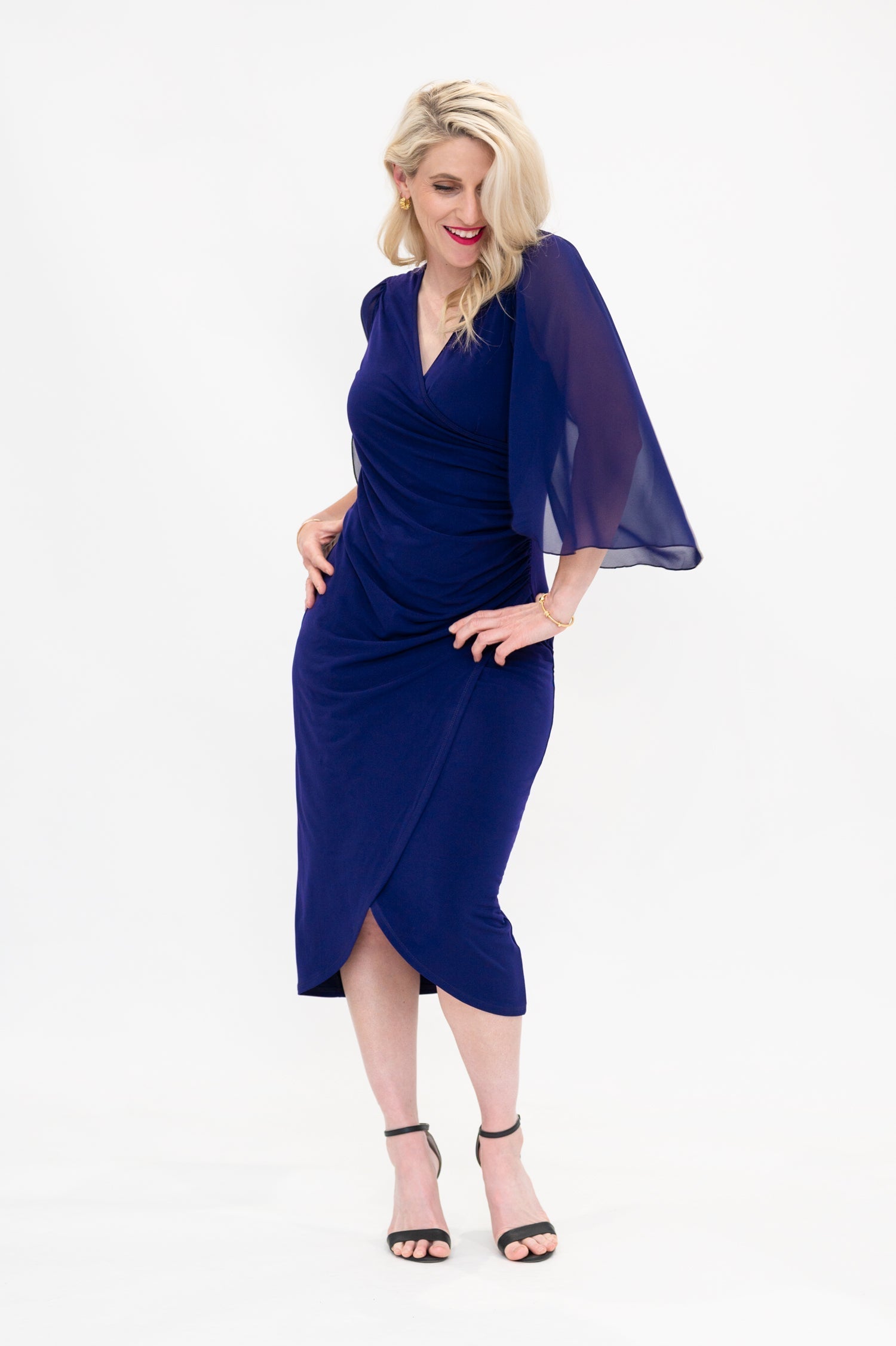 Female model dancing while wearing semi formal cocktail knee length faux wrap dress with tulip hemline and flared chiffon sleeves in deep navy colour