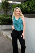 The female model wears a cowl neck top with no sleeves, the top is tucked into black pants. The print is small scale deep navy, white and teal geometric diamond shape pattern