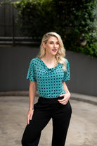 The female model wears a cowl neck top with flutter sleeves, the top is tucked into black pants. The print is small scale deep navy, white and teal geometric diamond shape pattern