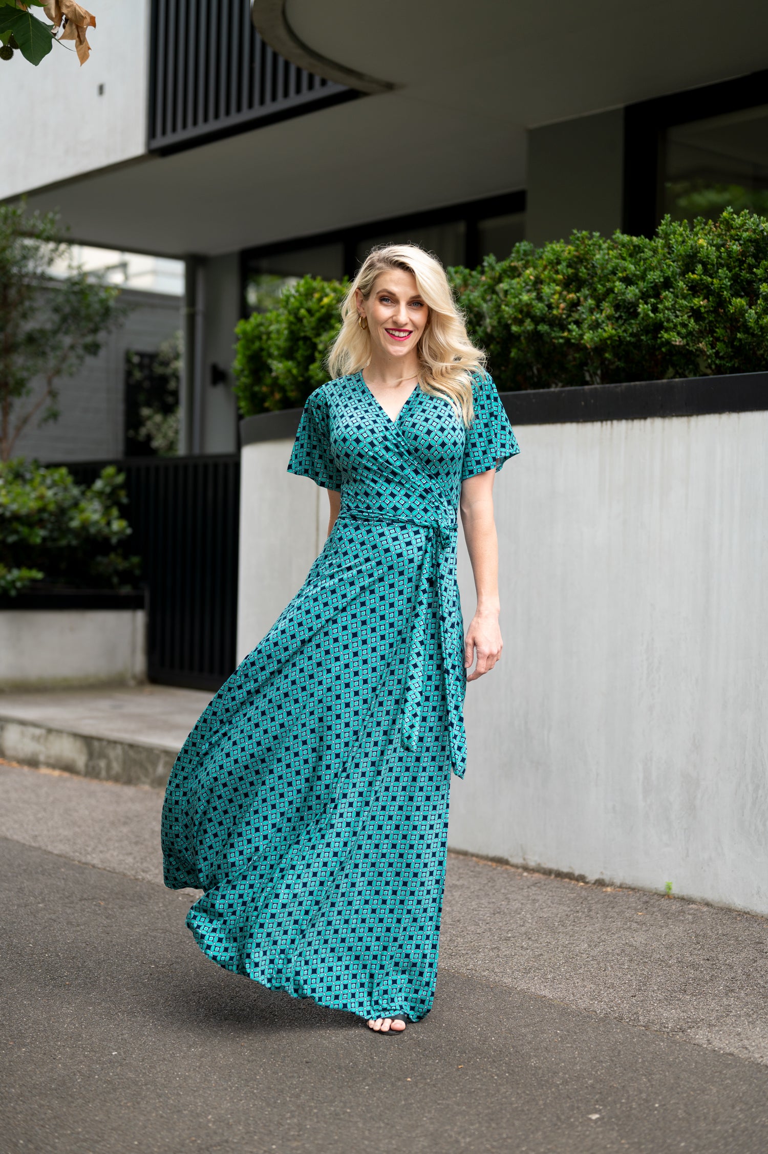 The female model wears a wrap dress in maxi length with flutter sleeves. The print is a small-scale deep navy, white and teal geometric diamond shape pattern. Front view